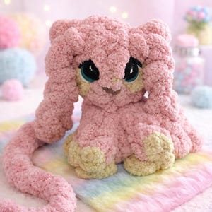 Puede incluir: Un peluche rosa hecho a mano con forma de cachorro de león, con ojos grandes y expresivos. El peluche tiene una textura esponjosa y está sentado sobre una superficie de colores arcoíris. Las patas y el interior de las orejas son de color amarillo claro.