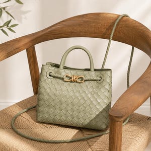 Pu&ograve; includere: Una borsa verde salvia con un design intrecciato &egrave; appoggiata su una sedia di legno. La borsa ha un manico superiore e una lunga tracolla. Una chiusura color oro adorna la parte anteriore. La sedia &egrave; in legno e materiale intrecciato.