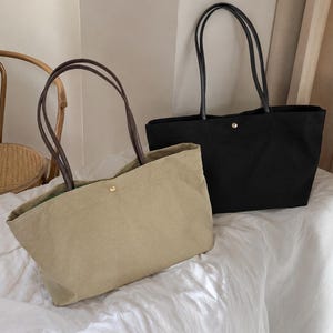 May include: Two rectangular tote bags, one in a light olive green and one in black, both featuring long dark brown shoulder straps and a single gold-toned metal snap button closure at the top center of the bag opening for secure storage.