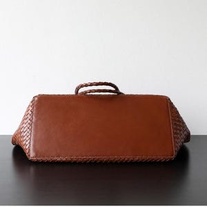 Op de afbeelding: Bruine leren tote bag met een geweven ontwerp aan de zijkanten en een gladde, rechthoekige basis. De tas heeft twee gevlochten handvatten en staat tegen een witte achtergrond.