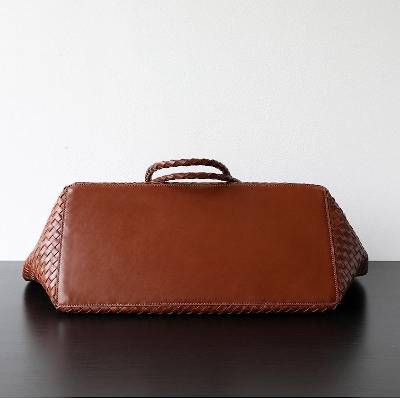 Op de afbeelding: Bruine leren tote bag met een geweven ontwerp aan de zijkanten en een gladde, rechthoekige basis. De tas heeft twee gevlochten handvatten en staat tegen een witte achtergrond.