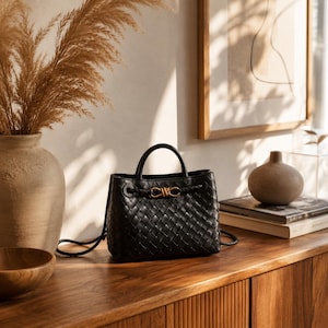 Pu&ograve; includere: Borsa a mano in pelle nera intrecciata con manico superiore e chiusura color oro. La borsa ha una forma strutturata e un dettaglio intrecciato sui lati. &Egrave; appoggiata su una superficie di legno con altri oggetti decorativi.
