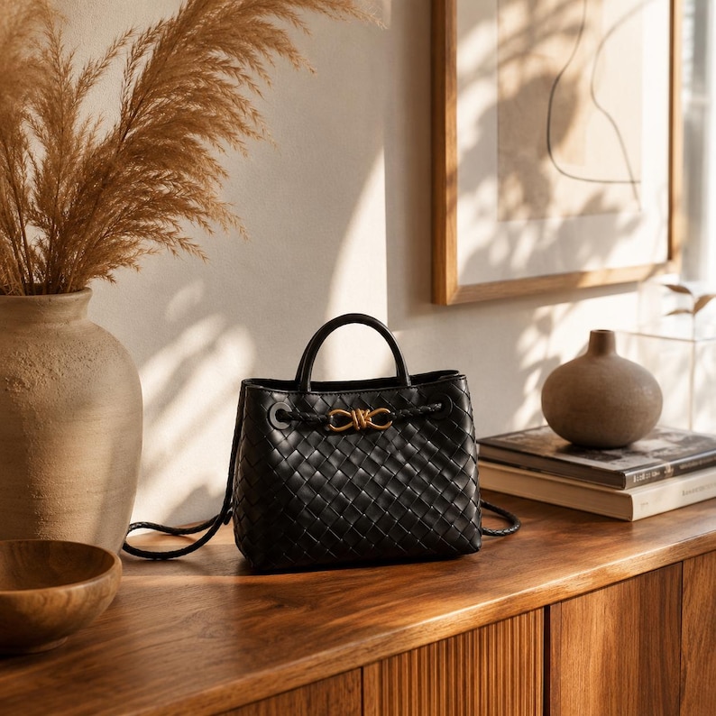 Pu&ograve; includere: Borsa a mano in pelle nera intrecciata con manico superiore e chiusura color oro. La borsa ha una forma strutturata e un dettaglio intrecciato sui lati. &Egrave; appoggiata su una superficie di legno con altri oggetti decorativi.