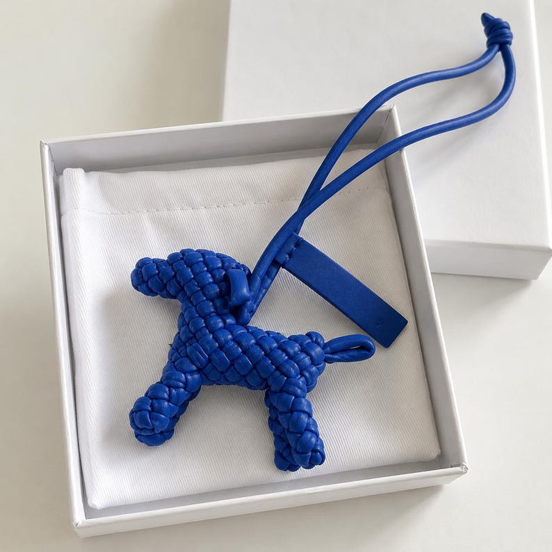 May include: A blue woven dog-shaped keychain with a matching blue strap and tag, presented in a white box with a white fabric lining. The dog charm has a textured, braided appearance.