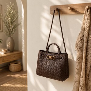 Pu&ograve; includere: Una borsa a mano in pelle intrecciata marrone scuro con chiusura color oro e tracolla intrecciata &egrave; appesa a un piolo di legno. La borsa ha una forma strutturata con un manico superiore e un accento decorativo.