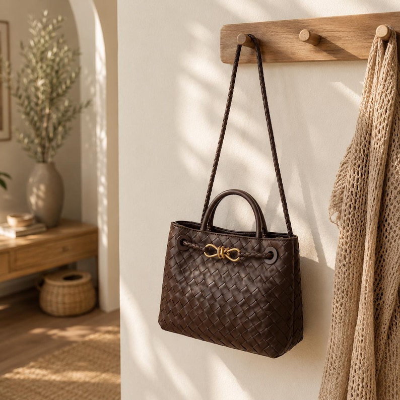 Pu&ograve; includere: Una borsa a mano in pelle intrecciata marrone scuro con chiusura color oro e tracolla intrecciata &egrave; appesa a un piolo di legno. La borsa ha una forma strutturata con un manico superiore e un accento decorativo.