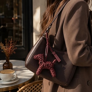 May include: A dark brown leather shoulder bag with a matching strap. A burgundy woven dog-shaped charm hangs from the strap. The bag is worn over a brown blazer.