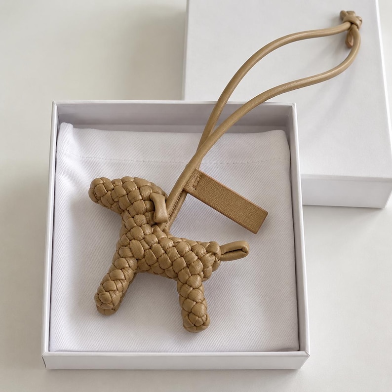 May include: A tan leather dog-shaped keychain with a woven texture, presented in a white box. The keychain has a loop for attachment and a rectangular tag. The dog's form is detailed, and the overall aesthetic is minimalist.