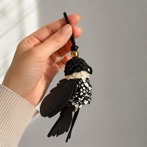 May include: A bird-shaped bag charm crafted from layered black and white leather pieces. The design features a gold-toned metal bead at the top, a black cord loop for attachment, and textured wing details. The body is composed of small, overlapping leather scales.