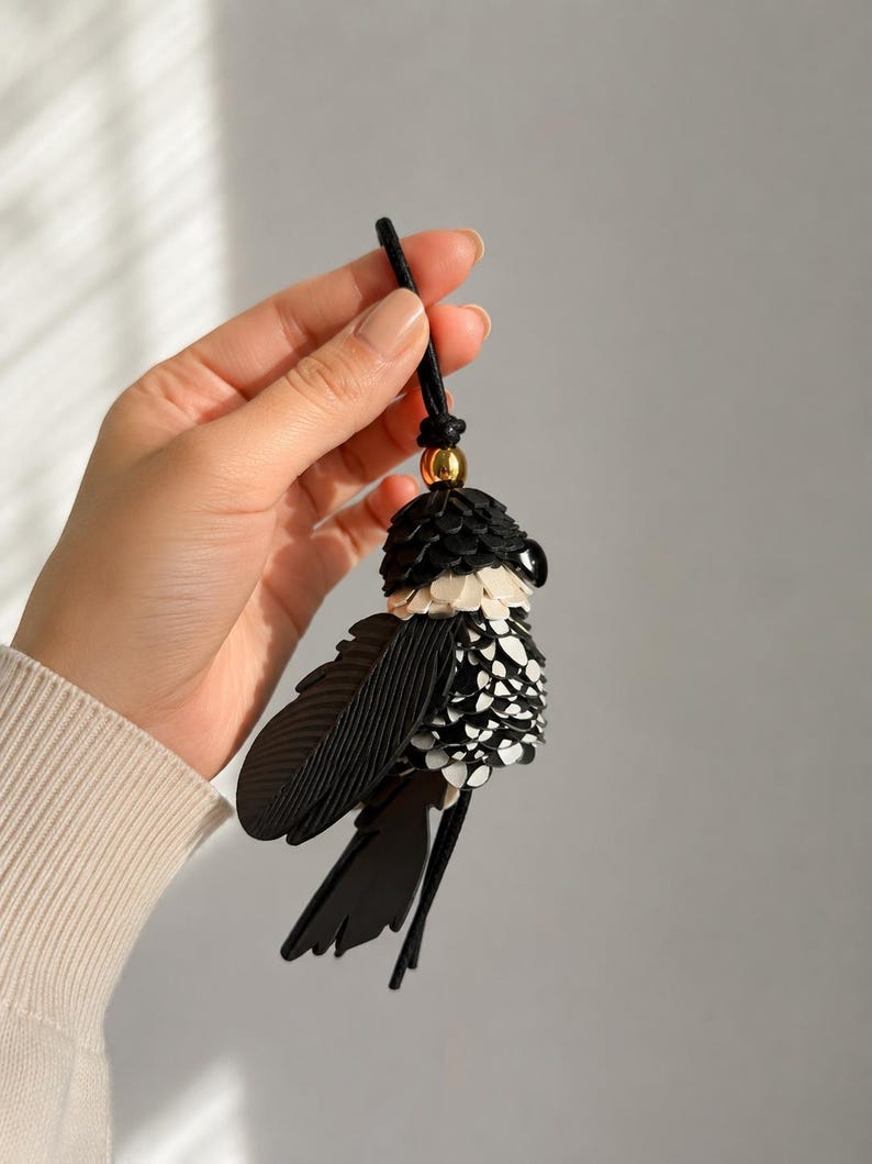 May include: A bird-shaped bag charm crafted from layered black and white leather pieces. The design features a gold-toned metal bead at the top, a black cord loop for attachment, and textured wing details. The body is composed of small, overlapping leather scales.