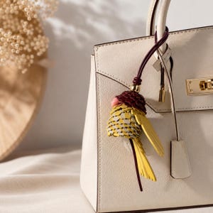 May include: A cream-colored leather handbag featuring a gold-toned metal clasp and a decorative bird-shaped charm. The charm is crafted from layered yellow, grey, and dark red leather pieces, suspended from a dark red cord attached to the handle.