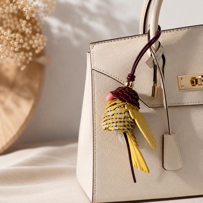 May include: A cream-colored leather handbag featuring a gold-toned metal clasp and a decorative bird-shaped charm. The charm is crafted from layered yellow, grey, and dark red leather pieces, suspended from a dark red cord attached to the handle.