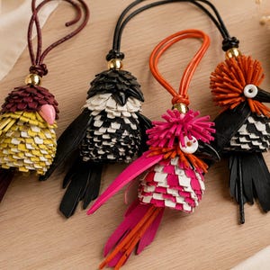 May include: Four bird-shaped bag charms crafted from layered leather strips. Each features a gold-tone bead, a hanging cord, and a unique color scheme including yellow, burgundy, black, white, orange, and pink with fringe-like tail feathers.
