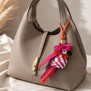 May include: A taupe-colored handbag with a textured surface, a single top handle, and a gold-toned metal clasp closure. Attached to the handle is a decorative bag charm shaped like a bird, featuring vibrant pink, white, and orange leather fringe.