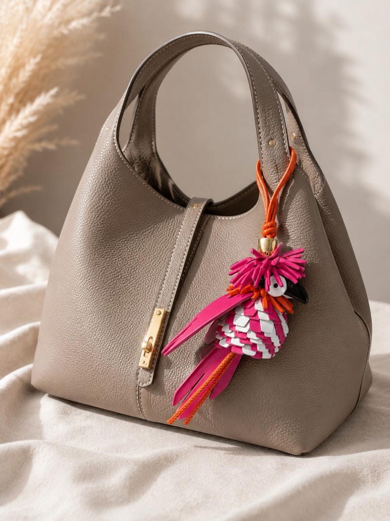 May include: A taupe-colored handbag with a textured surface, a single top handle, and a gold-toned metal clasp closure. Attached to the handle is a decorative bag charm shaped like a bird, featuring vibrant pink, white, and orange leather fringe.