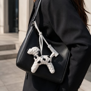 May include: Black leather shoulder bag with a white woven dog charm featuring black spots. The bag has a single strap and a white tassel. The dog charm is attached to the bag with a white cord.