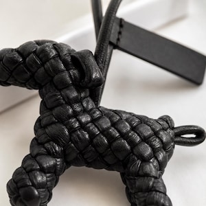 May include: A black leather dog-shaped keychain with a woven design. The keychain features a loop for attachment and a rectangular tag. The dog's body is intricately woven, showcasing a detailed texture.