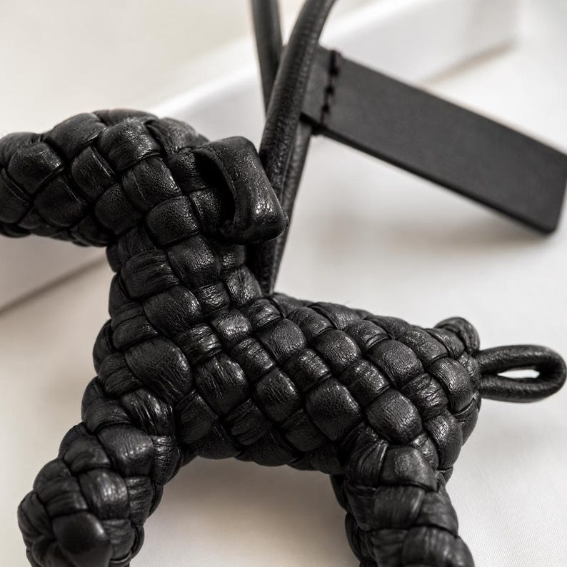May include: A black leather dog-shaped keychain with a woven design. The keychain features a loop for attachment and a rectangular tag. The dog's body is intricately woven, showcasing a detailed texture.
