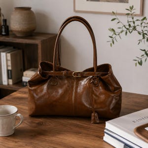 May include: A glossy brown leather handbag featuring a structured rectangular base, two long shoulder straps, and a thin horizontal belt detail across the top opening with a small gold-toned metal buckle and a dangling leather tab accent.