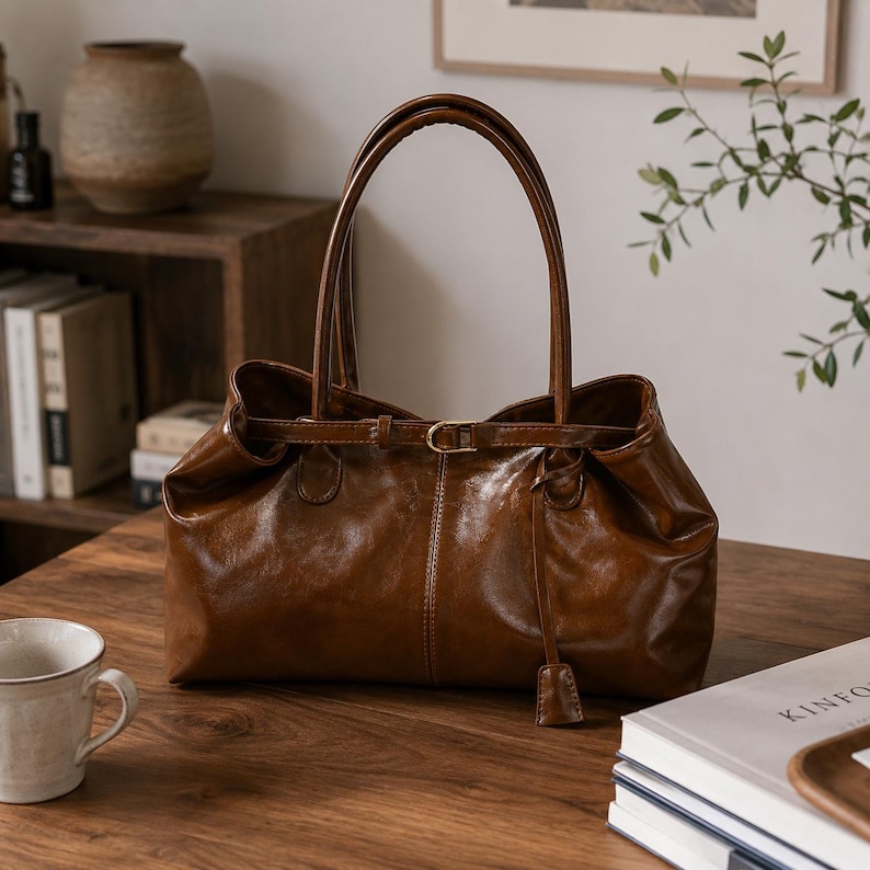 May include: A glossy brown leather handbag featuring a structured rectangular base, two long shoulder straps, and a thin horizontal belt detail across the top opening with a small gold-toned metal buckle and a dangling leather tab accent.