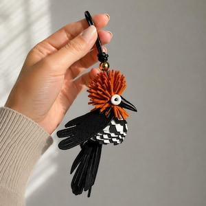 May include: A bird-shaped bag charm crafted from black, white, and burnt orange leather pieces. The bird features a prominent orange fringed crest, a sharp black beak, a white circular eye, black wings, and a layered black and white tail.
