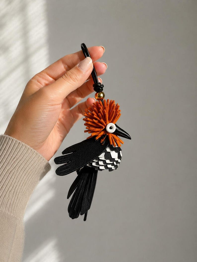 May include: A bird-shaped bag charm crafted from black, white, and burnt orange leather pieces. The bird features a prominent orange fringed crest, a sharp black beak, a white circular eye, black wings, and a layered black and white tail.