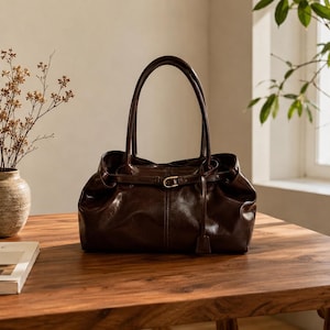 May include: A dark brown leather handbag with a smooth, glossy finish, featuring two tall shoulder straps and a thin horizontal belt detail across the front secured by a small gold-toned metal buckle and a dangling leather tab.