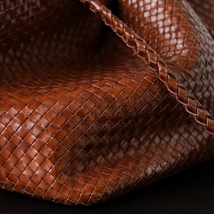 Op de afbeelding: Close-up van een bruine leren tote bag. De tas heeft een geweven ontwerp en een gevlochten riem. Het leer heeft een rijke, warme tint en de tas lijkt ruim en goed gemaakt. De tas staat op een donkere ondergrond.