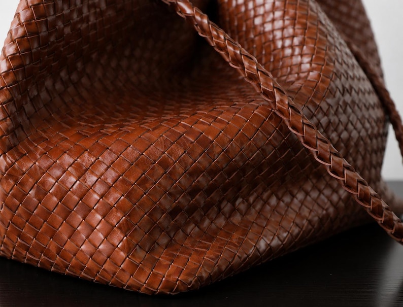 Op de afbeelding: Close-up van een bruine leren tote bag. De tas heeft een geweven ontwerp en een gevlochten riem. Het leer heeft een rijke, warme tint en de tas lijkt ruim en goed gemaakt. De tas staat op een donkere ondergrond.
