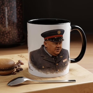 Peut inclure: Mug en céramique blanche avec intérieur et anse noirs. La tasse présente une illustration détaillée d'un homme en uniforme et chapeau sombres. Une cuillère et une pâtisserie sont sur une surface en bois.