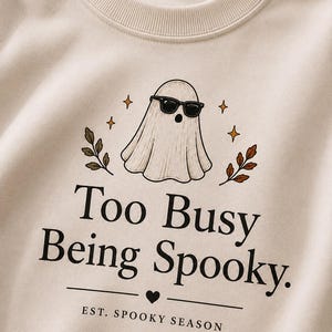 Puede incluir: Una sudadera beige de cuello redondo con un gráfico de un fantasma con gafas de sol negras, rodeado de pequeñas estrellas y hojas de otoño. El texto dice "Too Busy Being Spooky." y "EST. SPOOKY SEASON" con un pequeño icono de corazón.