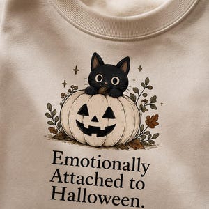 Puede incluir: Una sudadera beige de cuello redondo con un gráfico de un gato negro asomándose por una calabaza de Halloween rodeada de hojas. El texto dice "Emotionally Attached to Halloween." y "AUTUMN SOCIETY" con un pequeño corazón debajo.