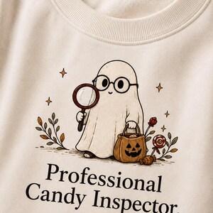 Puede incluir: Una sudadera de cuello redondo color crema con un gráfico de un fantasma con gafas redondas, sosteniendo una lupa y un cubo de dulces de calabaza. El texto dice Professional Candy Inspector. EST. OCTOBER.