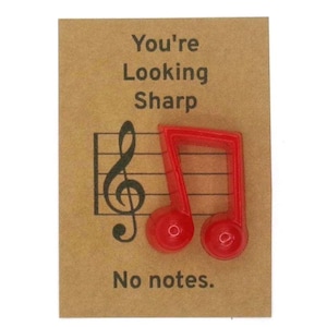 Puede incluir: Un objeto rojo en forma de nota musical sobre una tarjeta marrón con el texto "You're Looking Sharp" y "No notes." También hay una clave de sol y un pentagrama. El objeto es de un rojo brillante y sólido.