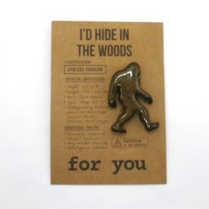 Op de afbeelding: Een donkerbruine, glanzende Bigfoot-vormige emaille speld bevestigd op een rechthoekige beige kaart. De kaart bevat de tekst I'D HIDE IN THE WOODS for you, samen met gedrukte details over classificatie, fysieke beschrijving en gedragskenmerken.