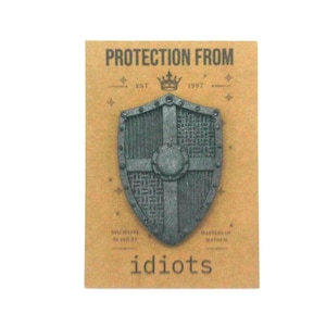 May include: A dark gray shield-shaped pin attached to a tan rectangular card. The card features the text PROTECTION FROM, EST. 1997, DISCIPLINE IN DOUBT, MASTERS OF MAYHEM, and idiots. The shield has a central cross and textured quadrants.
