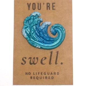 May include: A blue and teal wave-shaped pin with white accents, mounted on a brown card. The card reads "YOU'RE swell." and "NO LIFEGUARD REQUIRED."