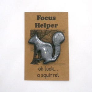 Puede incluir: Un broche con forma de ardilla gris sujeto a una tarjeta rectangular de color canela. La tarjeta presenta el texto Focus Helper en la parte superior y oh look... a squirrel en la parte inferior, con una silueta de árbol oscura impresa.