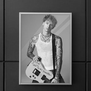 Op de afbeelding: Zwart-wit ingelijste print met een persoon met tatoeages, gekleed in een witte tanktop en met een gitaar. De gitaar heeft de tekst "I'M SELLING TICKETS TO MY DOWNFALL". De afbeelding wordt weergegeven op een donkergrijze muur.