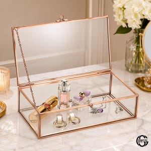 May include: A rectangular glass display box with a rose gold metal frame and a hinged lid held open by a thin chain. The interior contains a gold lipstick tube, a small perfume bottle, a pair of hoop earrings, and a delicate silver necklace.