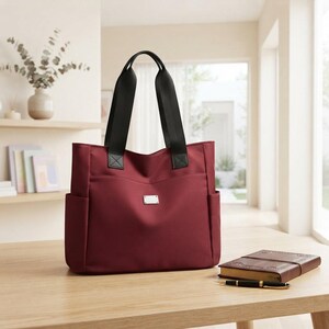 Puede incluir: Un bolso tote de color burdeos intenso con correas de hombro de nailon negro, una placa metálica rectangular de tono plateado en la parte delantera y bolsillos laterales. El bolso presenta un diseño estructurado con cierre de cremallera.