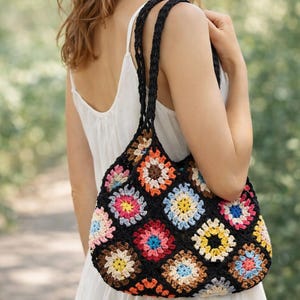Op de afbeelding: Een zwarte gehaakte tote bag met een kleurrijk bloemenpatroon. De tas heeft een reeks gehaakte vierkanten in verschillende kleuren, waaronder roze, blauw, geel en bruin. De tas heeft zwarte gevlochten handvatten.
