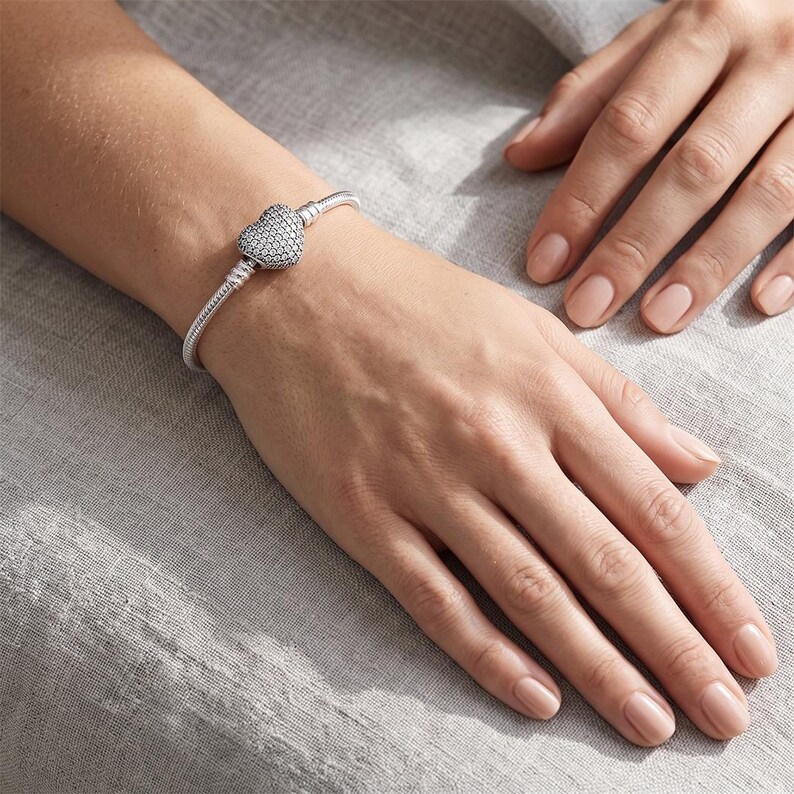 May include: A silver-toned snake chain bracelet featuring a heart-shaped clasp encrusted with numerous small, clear, sparkling stones. The bracelet is designed with a flexible, rounded metal link structure and a secure, prominent heart-shaped closure.