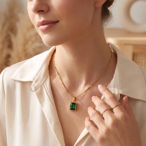 Op de afbeelding: Een goudkleurige ketting met een rechthoekige smaragdgroene hanger aan een fijne kralenketting. De hanger heeft een glad, gefacetteerd oppervlak en is gevat in een goudkleurige metalen zetting, rustend op een lichtgekleurd overhemd.