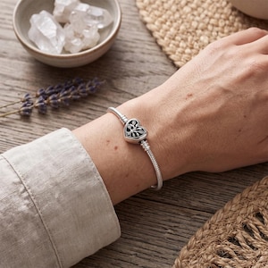 May include: A silver snake chain bracelet featuring a heart-shaped clasp with a detailed tree design. The clasp has a dark oxidized finish within the tree motif and small clear stones embedded in the branches, creating a contrast against the metal.