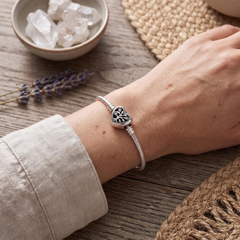 May include: A silver snake chain bracelet featuring a heart-shaped clasp with a detailed tree design. The clasp has a dark oxidized finish within the tree motif and small clear stones embedded in the branches, creating a contrast against the metal.