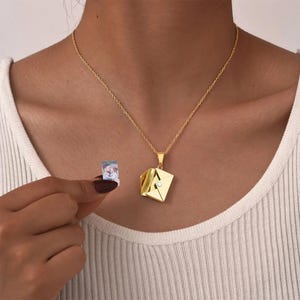 May include: A gold-toned necklace featuring a pendant shaped like a folded envelope. The envelope pendant has a small, clear, circular stone set on its front flap. A small, rectangular photograph showing two people is held separately near the pendant.
