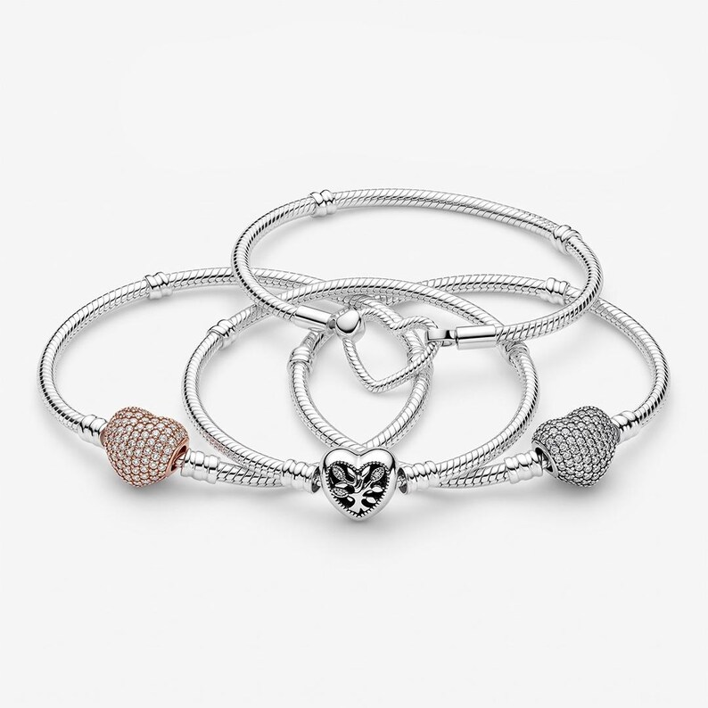 May include: Three silver snake chain bracelets featuring heart-shaped charms. One bracelet has a rose gold-toned heart charm with clear stones, one has a silver heart charm with a tree cutout, and one has a silver heart charm with clear stones.