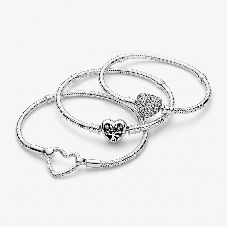 May include: Three silver snake chain bracelets. One features a heart-shaped clasp with a tree motif, another has a heart-shaped clasp covered in small clear stones, and the third has a simple open heart-shaped clasp. All are polished silver.