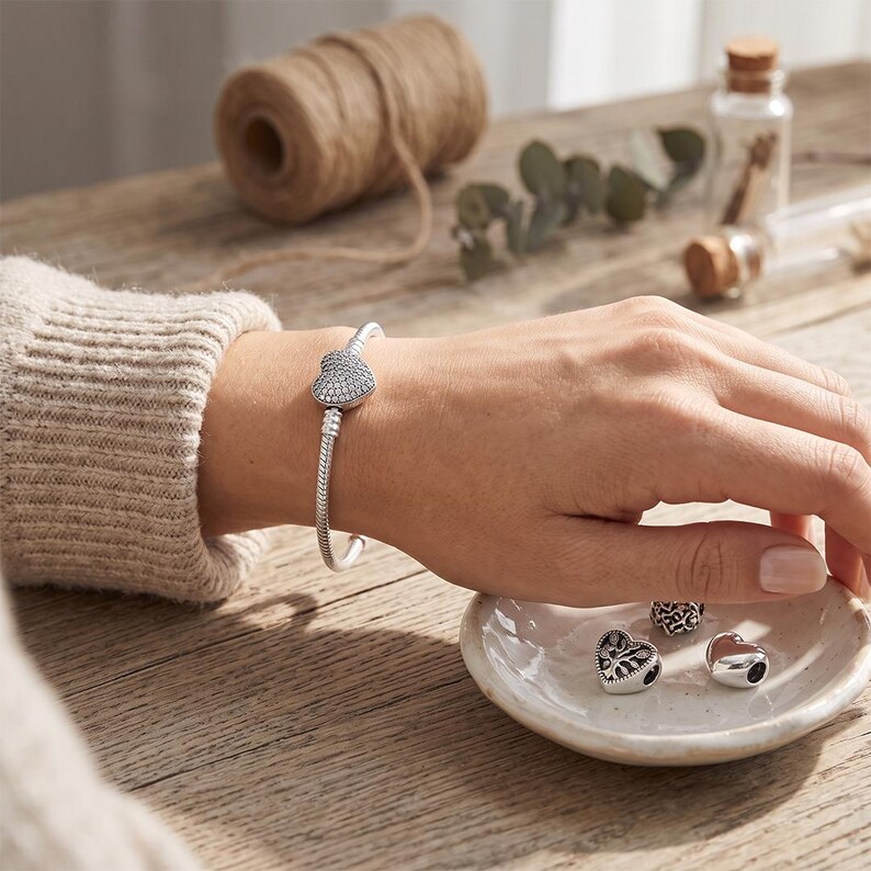 May include: A silver snake chain bracelet featuring a heart-shaped clasp covered in small clear stones. The bracelet is worn on a wrist, while three additional silver charms, including one heart-shaped charm with a tree design, rest nearby.